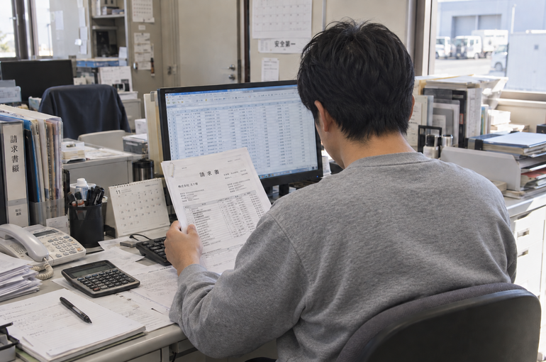 請求書とパソコンを確認しながら作業する男性の後ろ姿